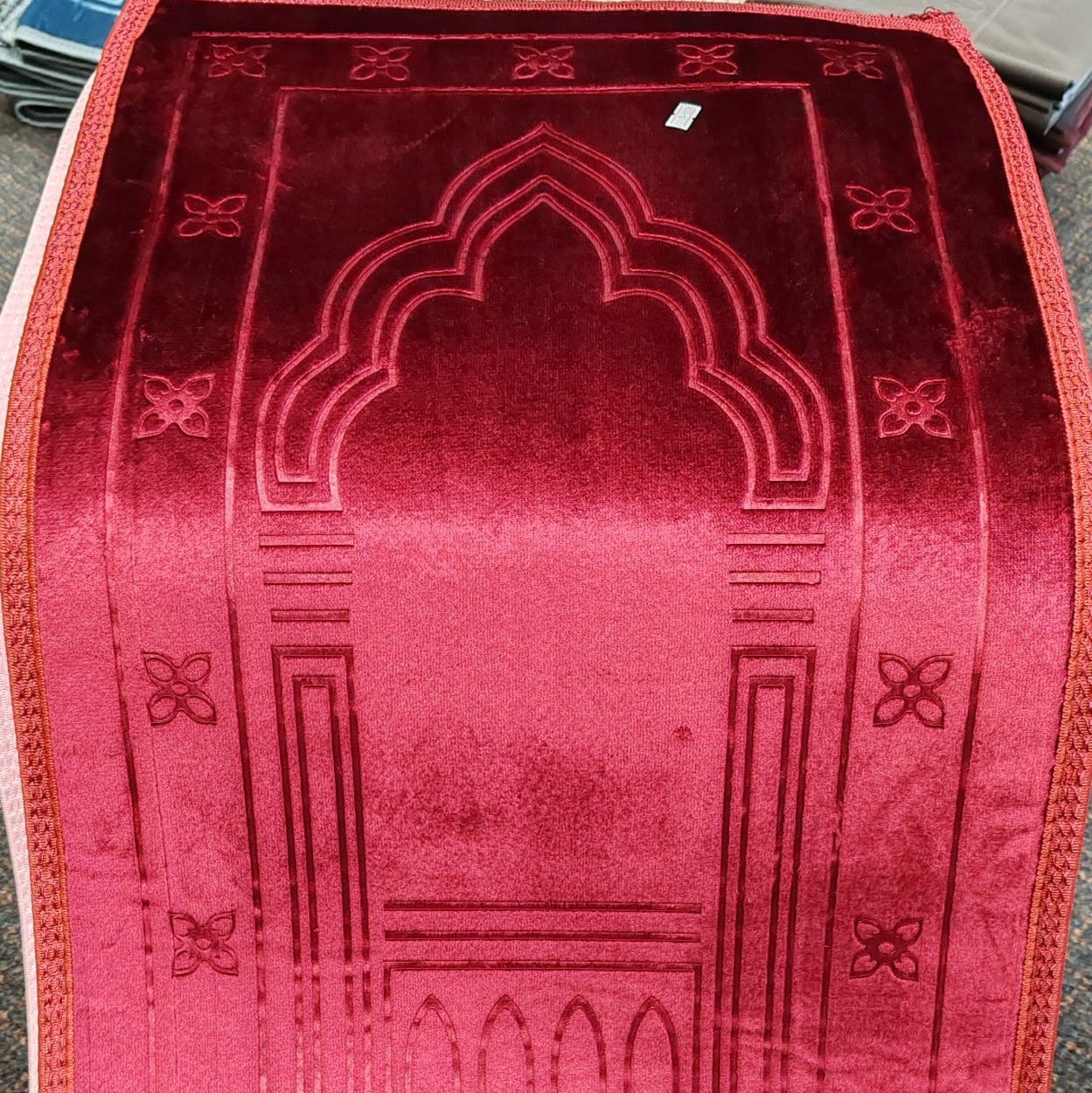 A red velvet prayer rug with an embossed mihrab arch design and geometric border patterns, laid flat on a dark surface.