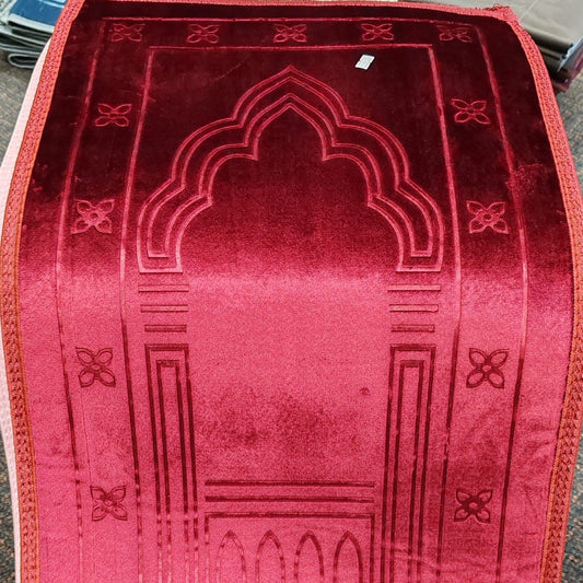A red velvet prayer rug with an embossed mihrab arch design and geometric border patterns, laid flat on a dark surface.